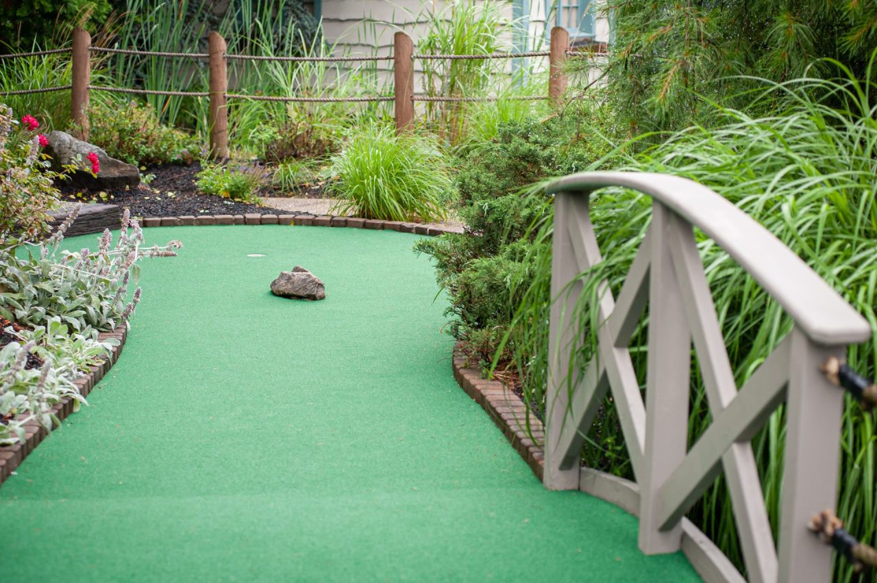 Miniature golf hole with green turf, a small rock obstacle, and brick edging next to a wooden fence and lush plants on both sides.