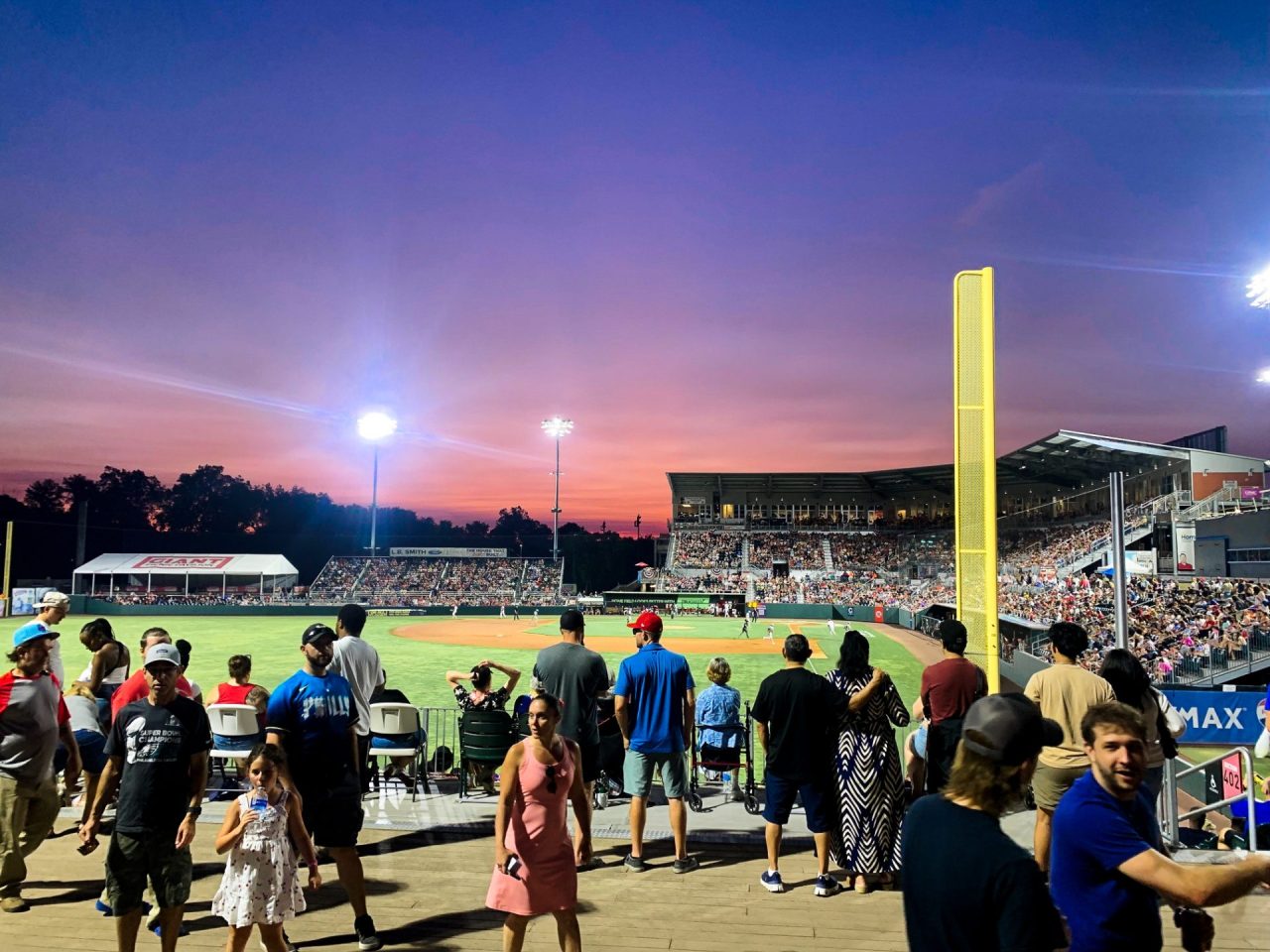 Crowd of baseball fans in a stadium at sunset, with a tall yellow foul post to the right and bright stadium lights.",