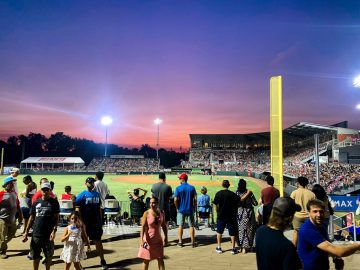 Crowd of baseball fans in a stadium at sunset, with a tall yellow foul post to the right and bright stadium lights.",