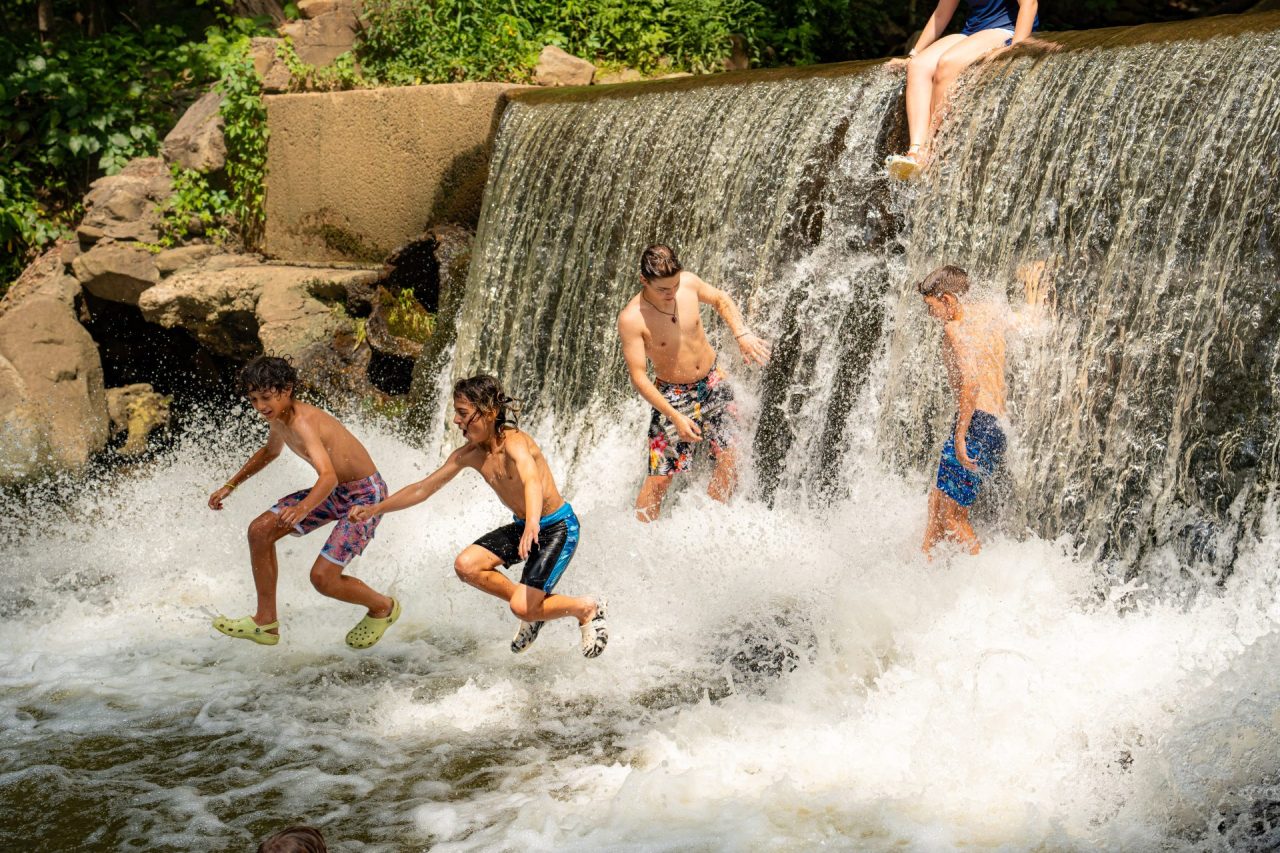 Several children in swim trunks and swimsuits jump from a rocky ledge into a splashy waterfall pool, surrounded by greenery.