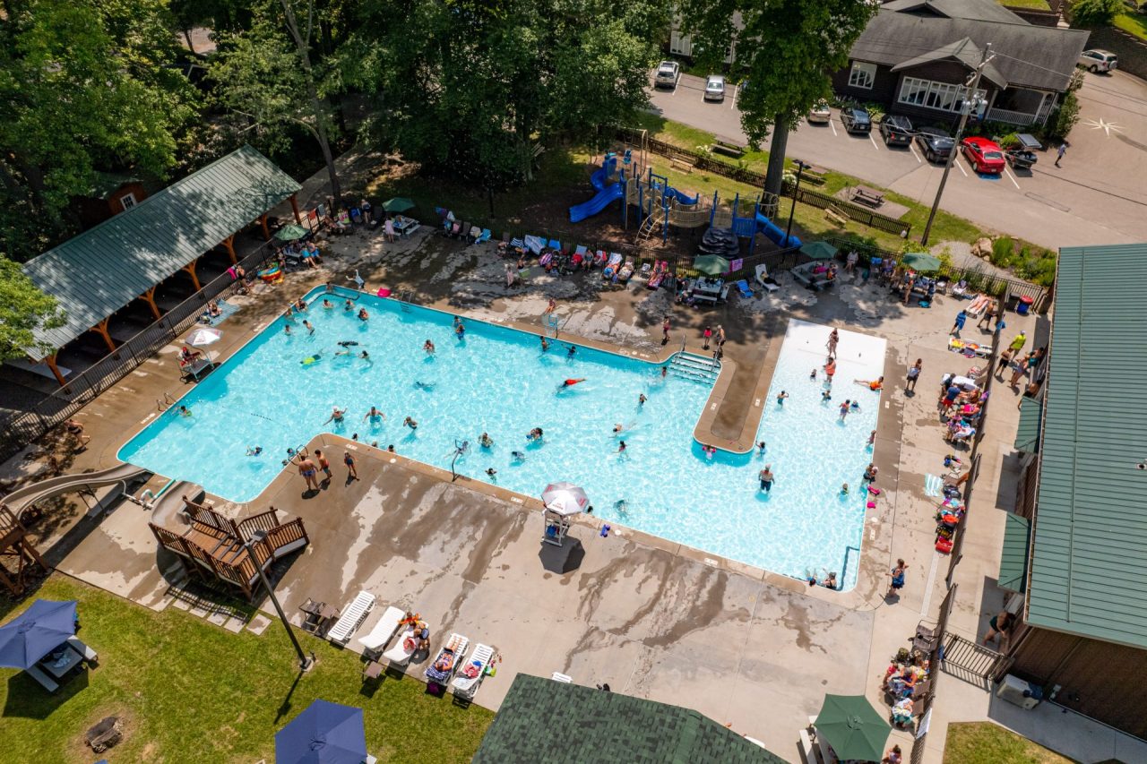 Aerial view of a busy outdoor pool area with swimmers, lounge chairs, umbrellas, and blue water.