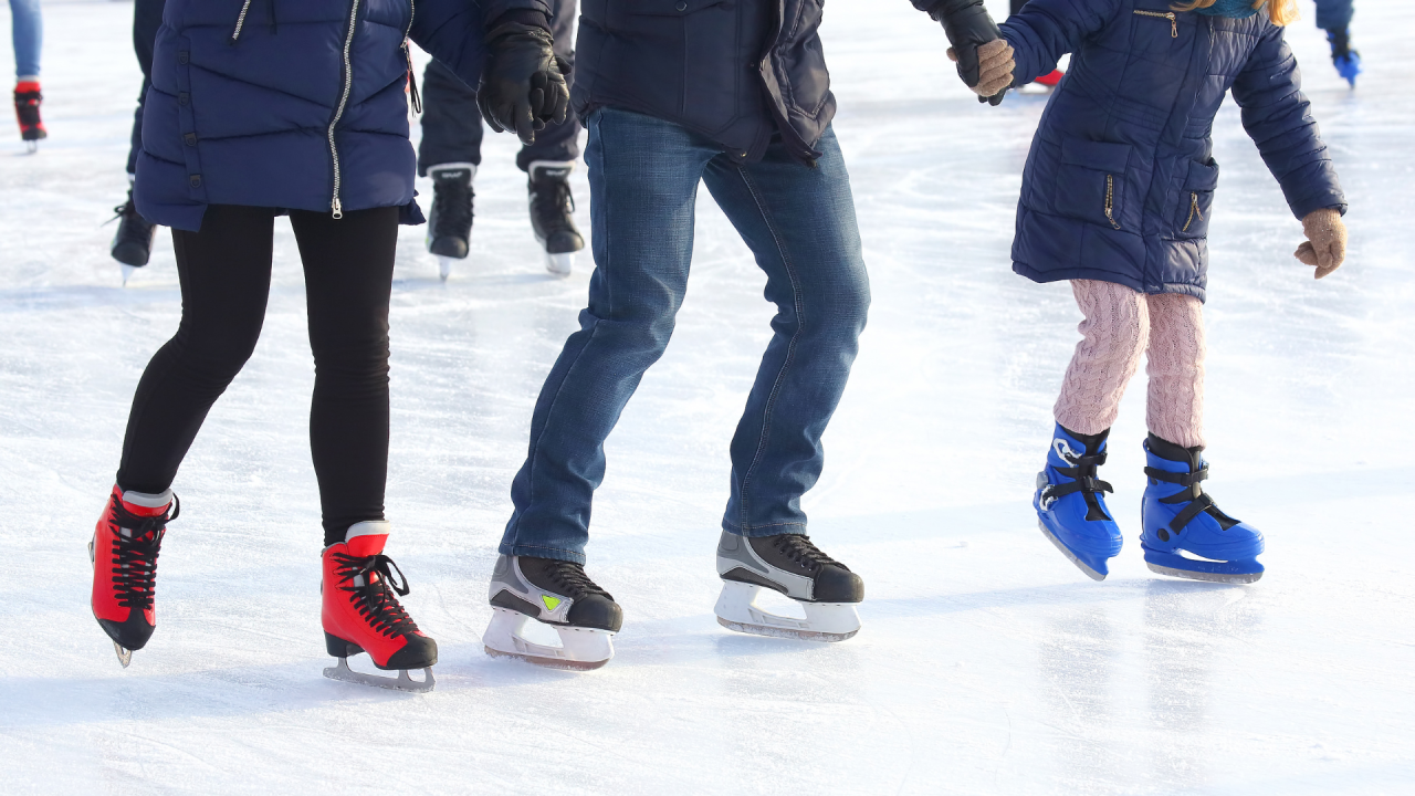 People ice skating on a rink, focus on colorful skates and winter jackets visible above the ice surface.