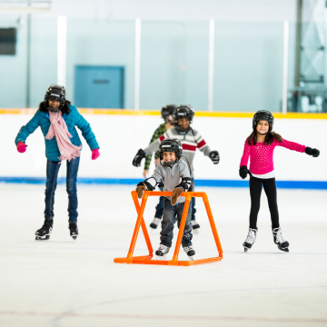 Child learning to ice skate with an orange support frame while other kids skate in a bright indoor rink, all wearing helmets and gloves.