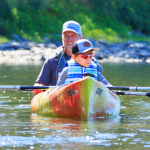 Grandfather and young boy paddling a colorful kayak on a calm river, wearing life jackets and caps.