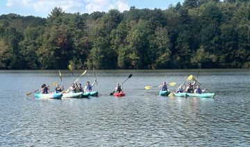 Group of people in colorful kayaks paddling on a calm lake with a tree-lined shore behind them