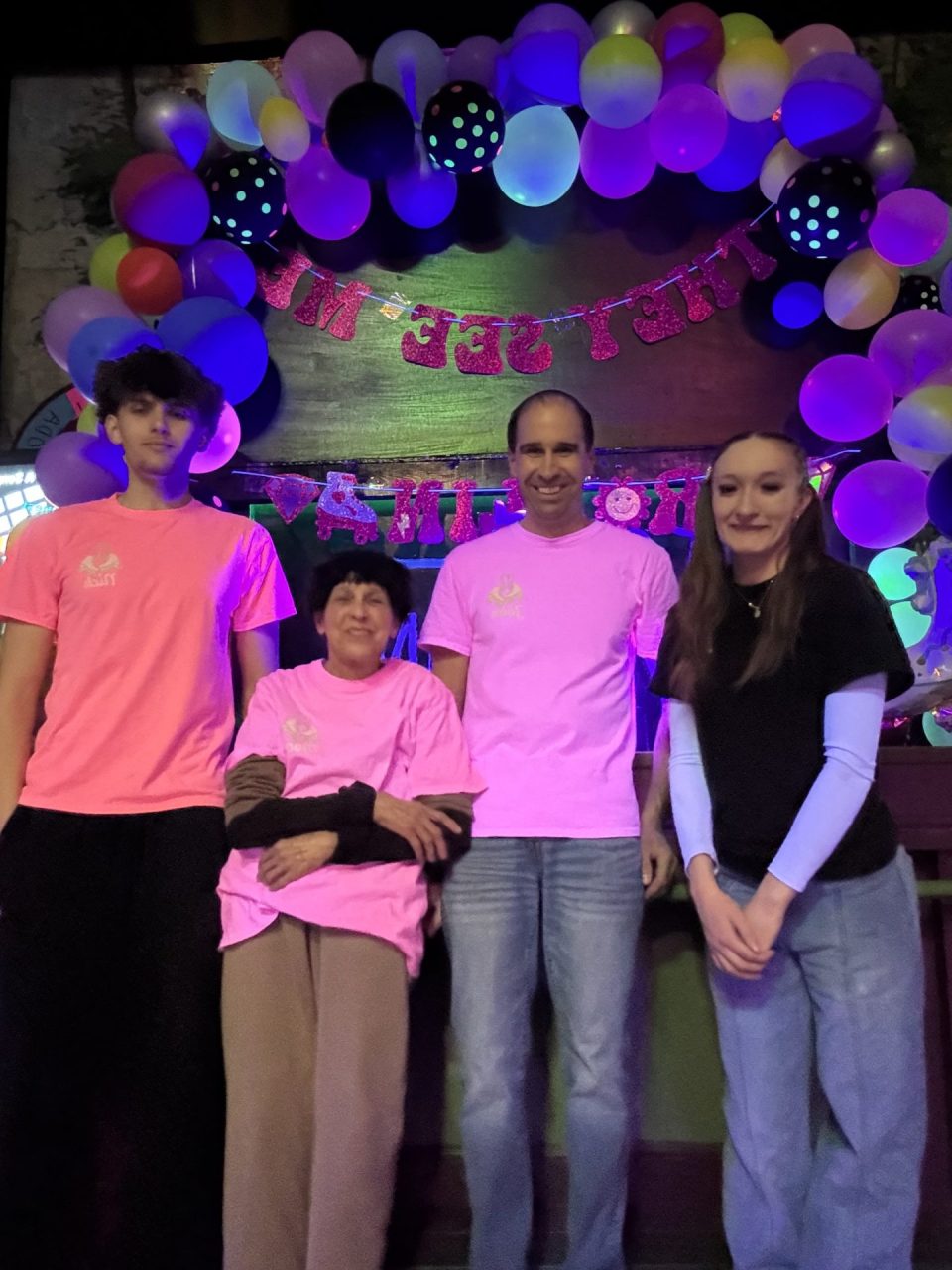 Four people pose for a birthday photo under a colorful balloon arch and pink glitter banner reading 'Happy Birthday.'