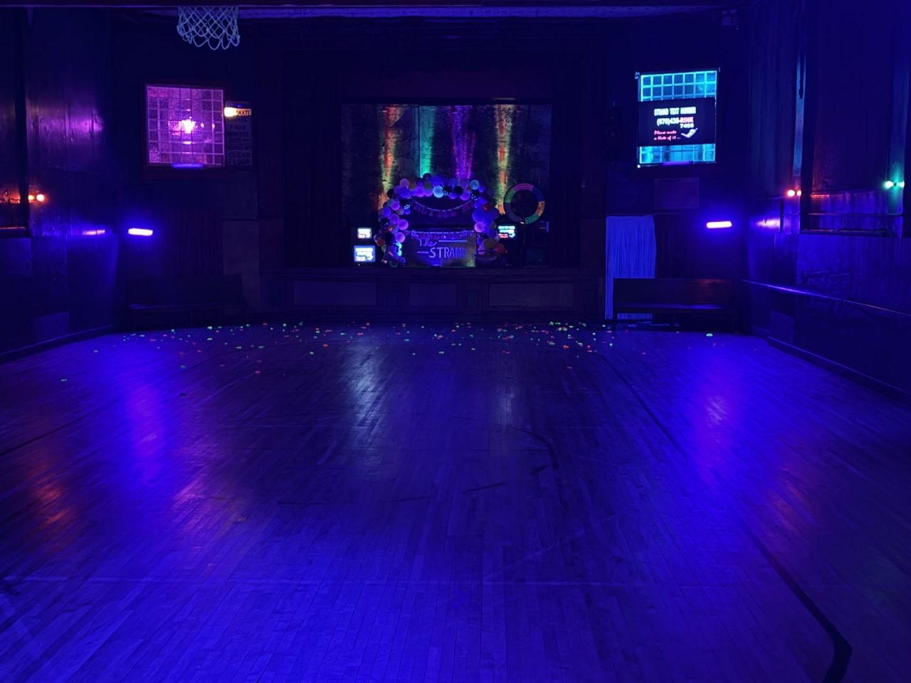 Dimly lit party venue with blue/purple neon lights, a decorated stage with a balloon arch, and confetti on the dance floor.