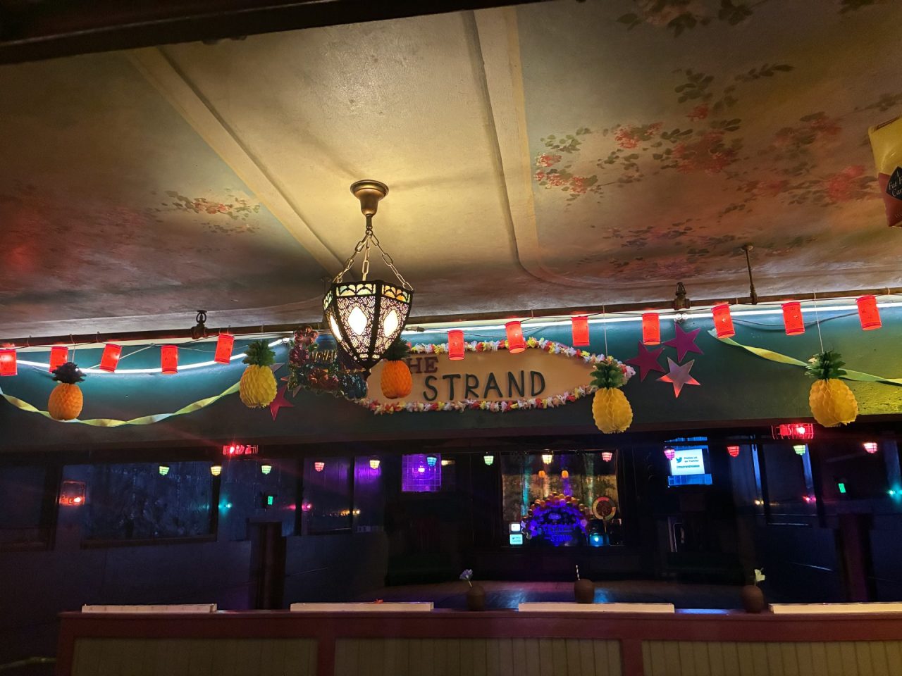 Festive indoor venue with a floral ceiling mural, ornate hanging lantern, and red lanterns plus pineapple decorations near a sign reading The Strand.