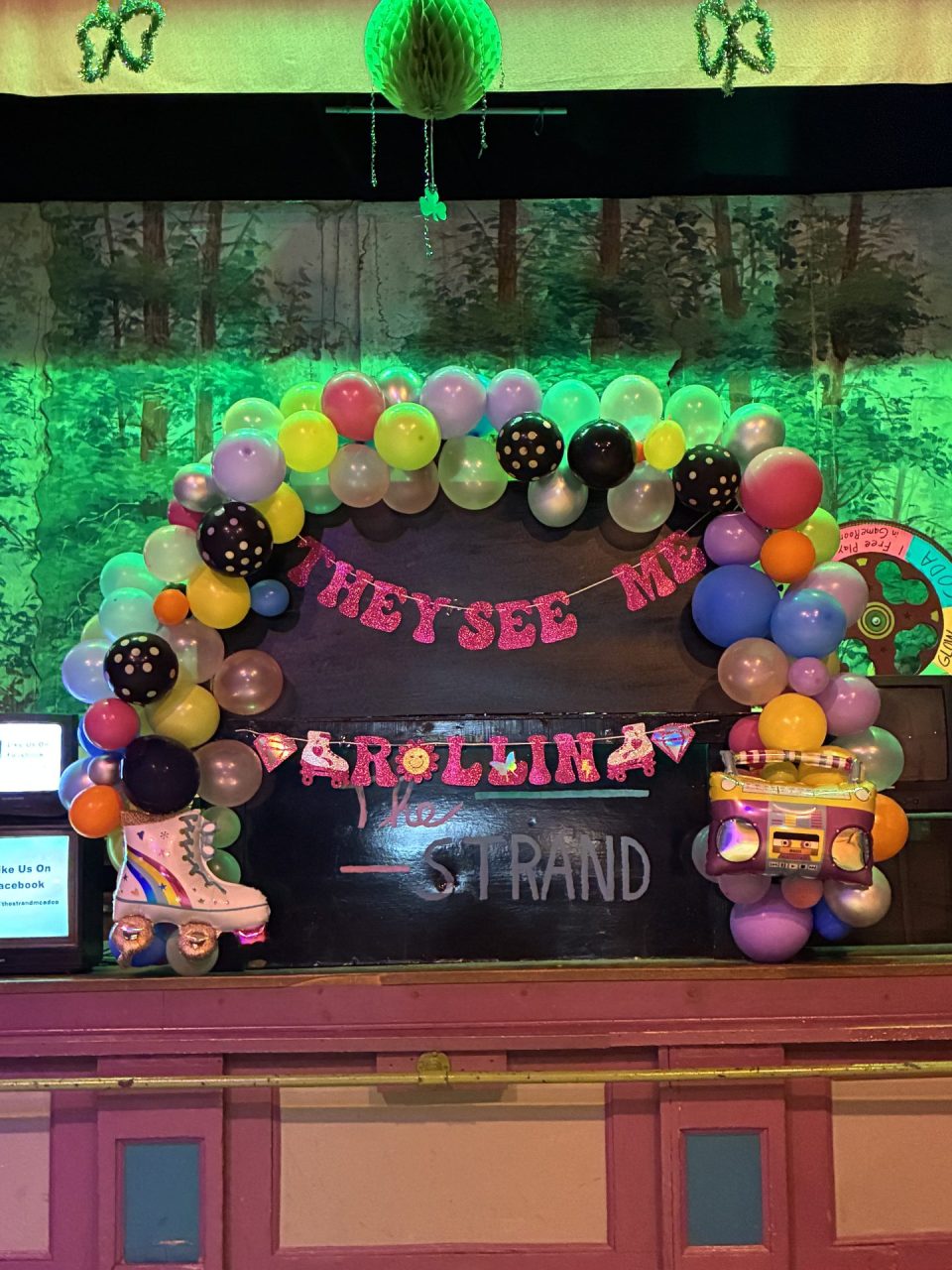 Colorful balloon arch decorating a stage with a forest mural backdrop and a pink glitter banner across the center of the scene.