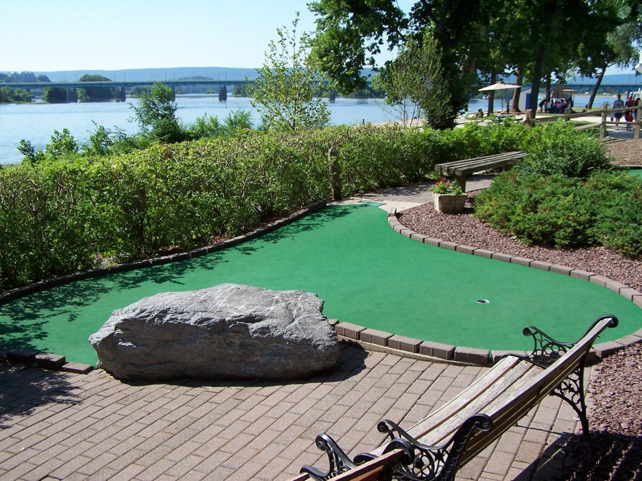 Riverside park with a green mini-golf putting green, a large rock, benches, and a distant bridge over the water.