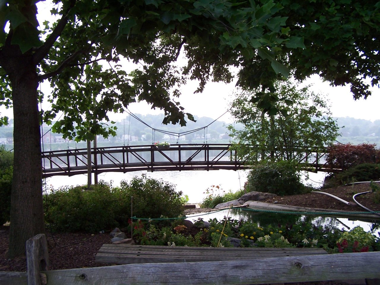 Wooden footbridge over a calm body of water, framed by trees and a garden with flowers; houses visible in the distance.