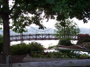 Wooden footbridge over a calm body of water, framed by trees and a garden with flowers; houses visible in the distance.