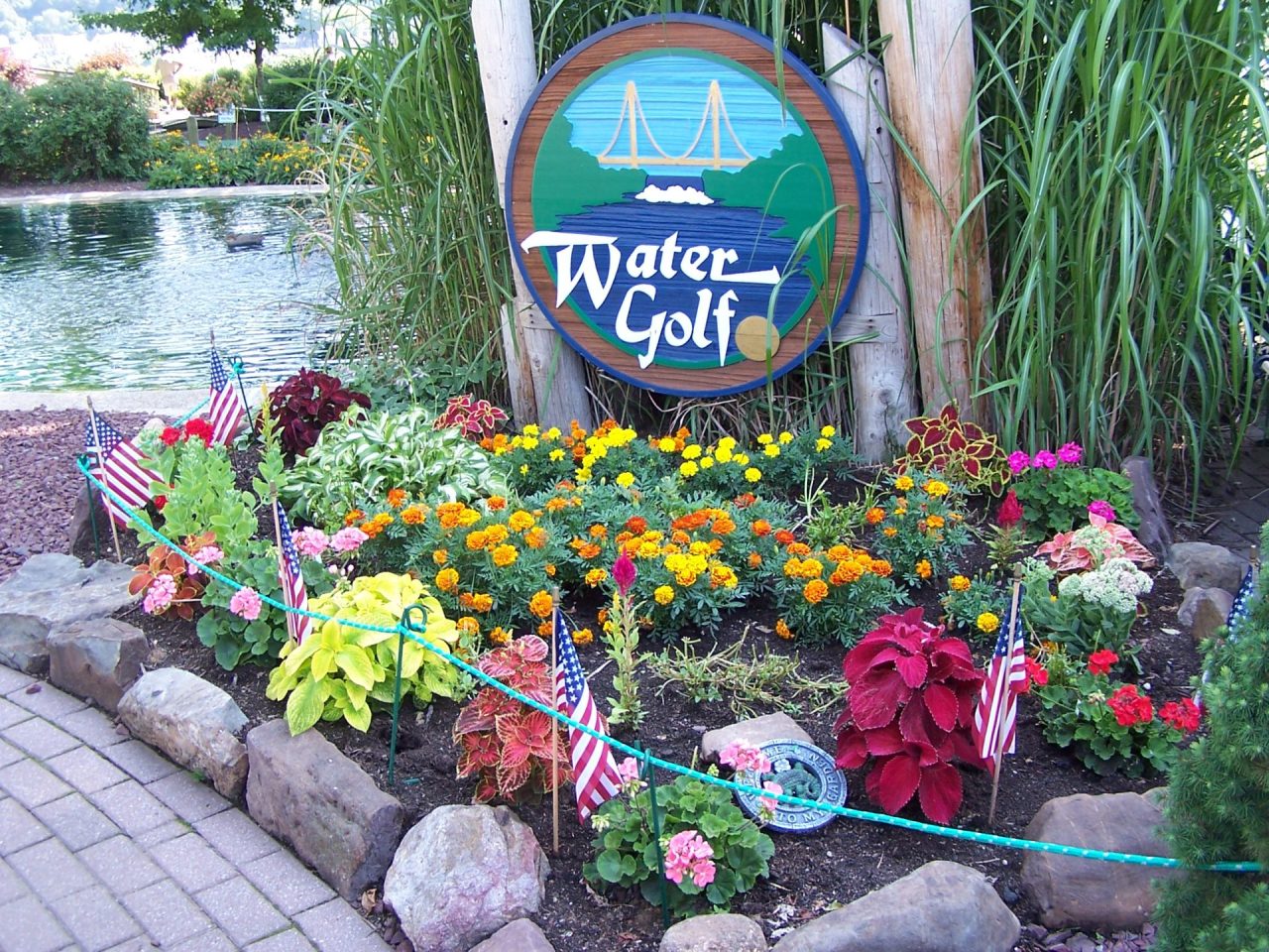 Circular Water Golf sign set in a flower bed with bright marigolds and red foliage, near a pond and decorative rocks with small American flags nearby.