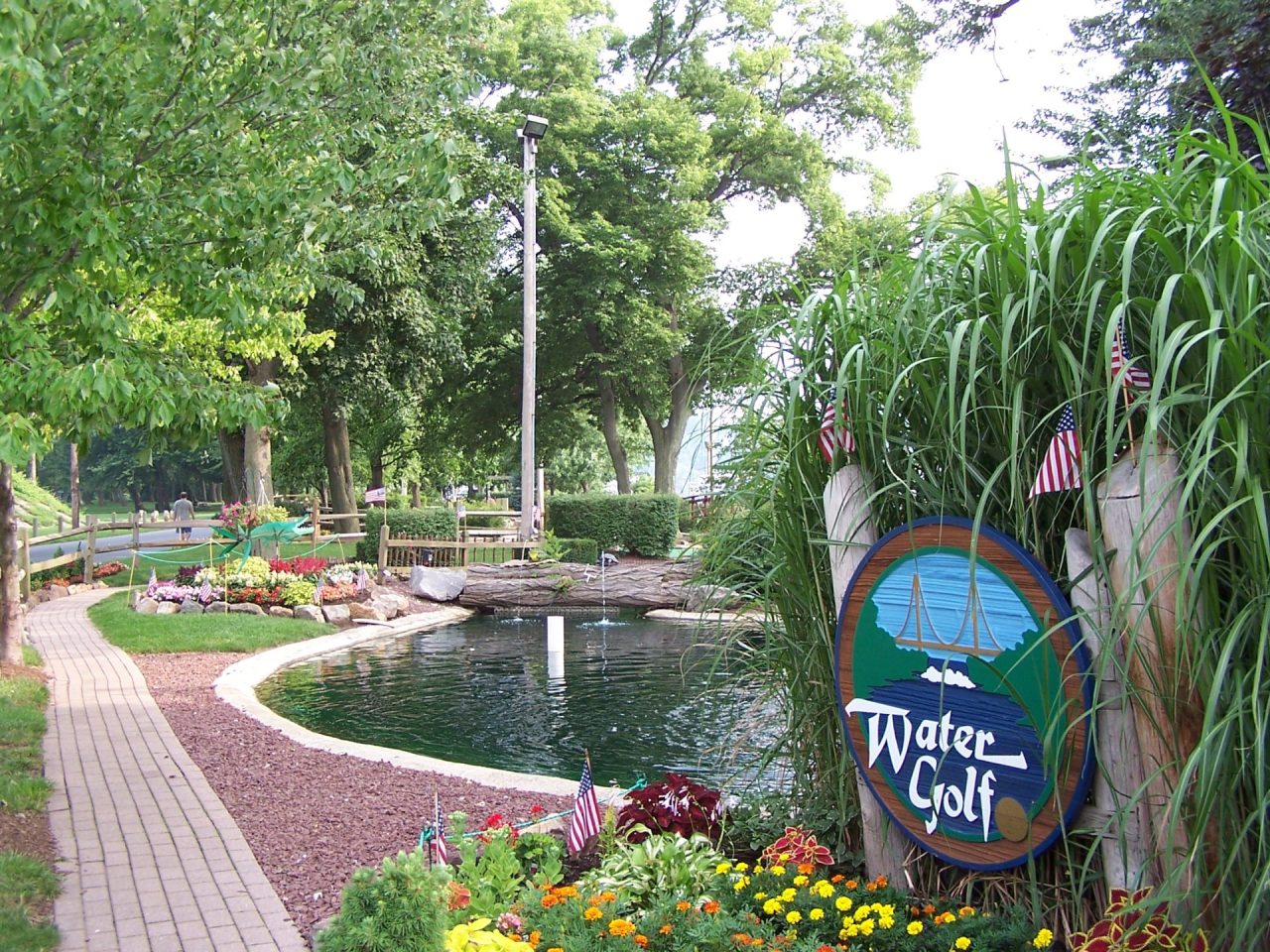 Park scene with a curved pond, brick path, and colorful flower beds beside tall green grasses and a round Water Golf sign.