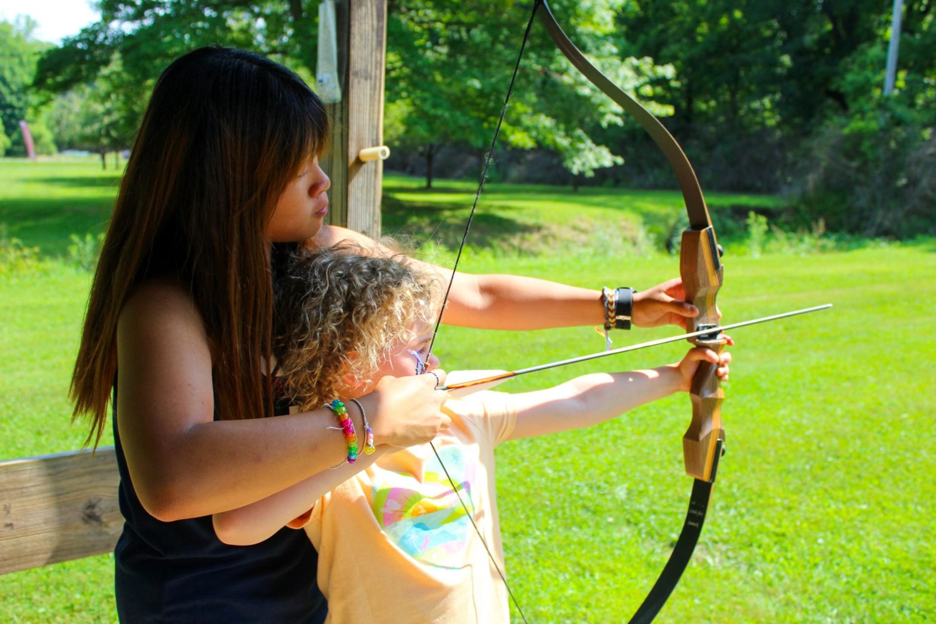 Two girls archery practice in a sunny park, one guiding the other as they aim a wooden recurve bow together.