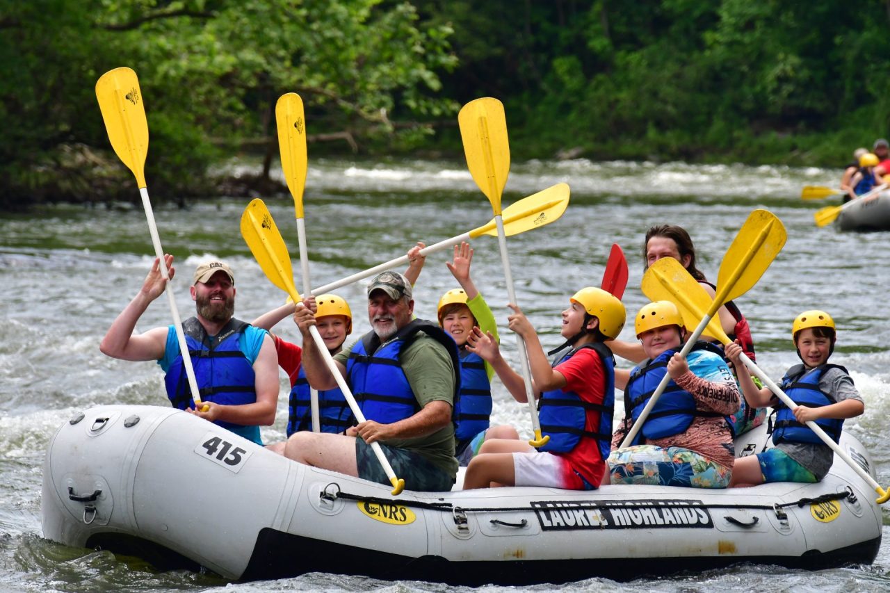 Group of people wearing yellow helmets and blue life jackets rafting on a gray inflatable boat with yellow paddles on a whitewater river, green trees in background.