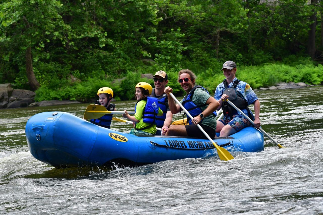 Group of five people whitewater rafting in a blue inflatable boat, wearing life jackets and helmets on a river with green foliage