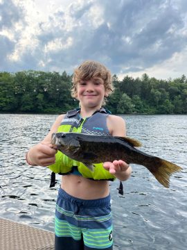 Young boy in a life jacket proudly holding a fish on a lakeside dock with a forested shore behind him.