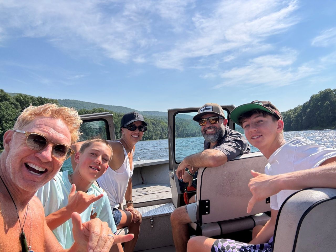 Group of friends and family taking a sunny boat selfie on a lake with hills in the background.