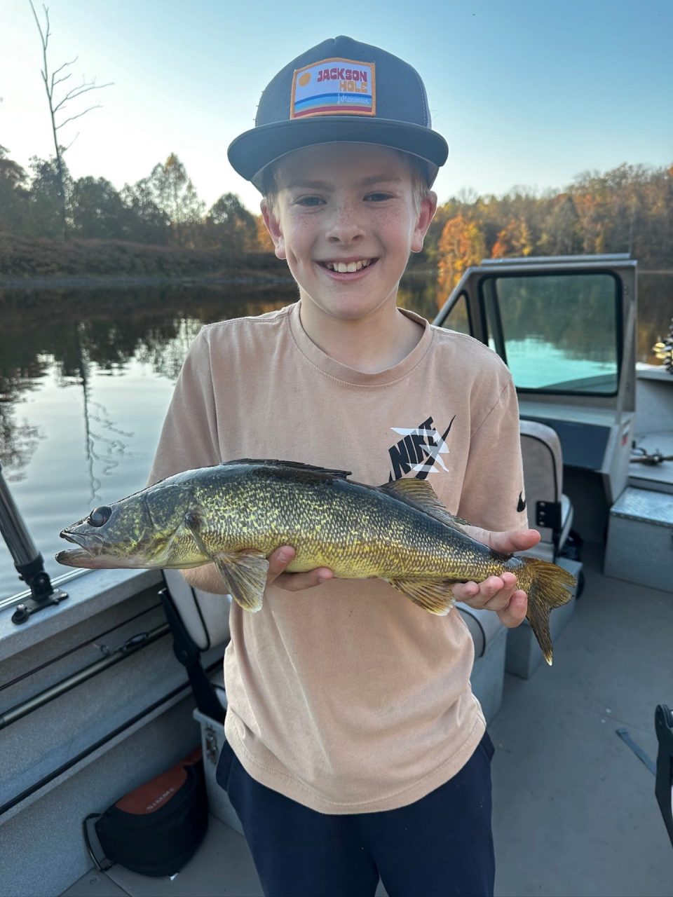 Boy on a boat proudly holds a large walleye fish with a calm lake in the background.