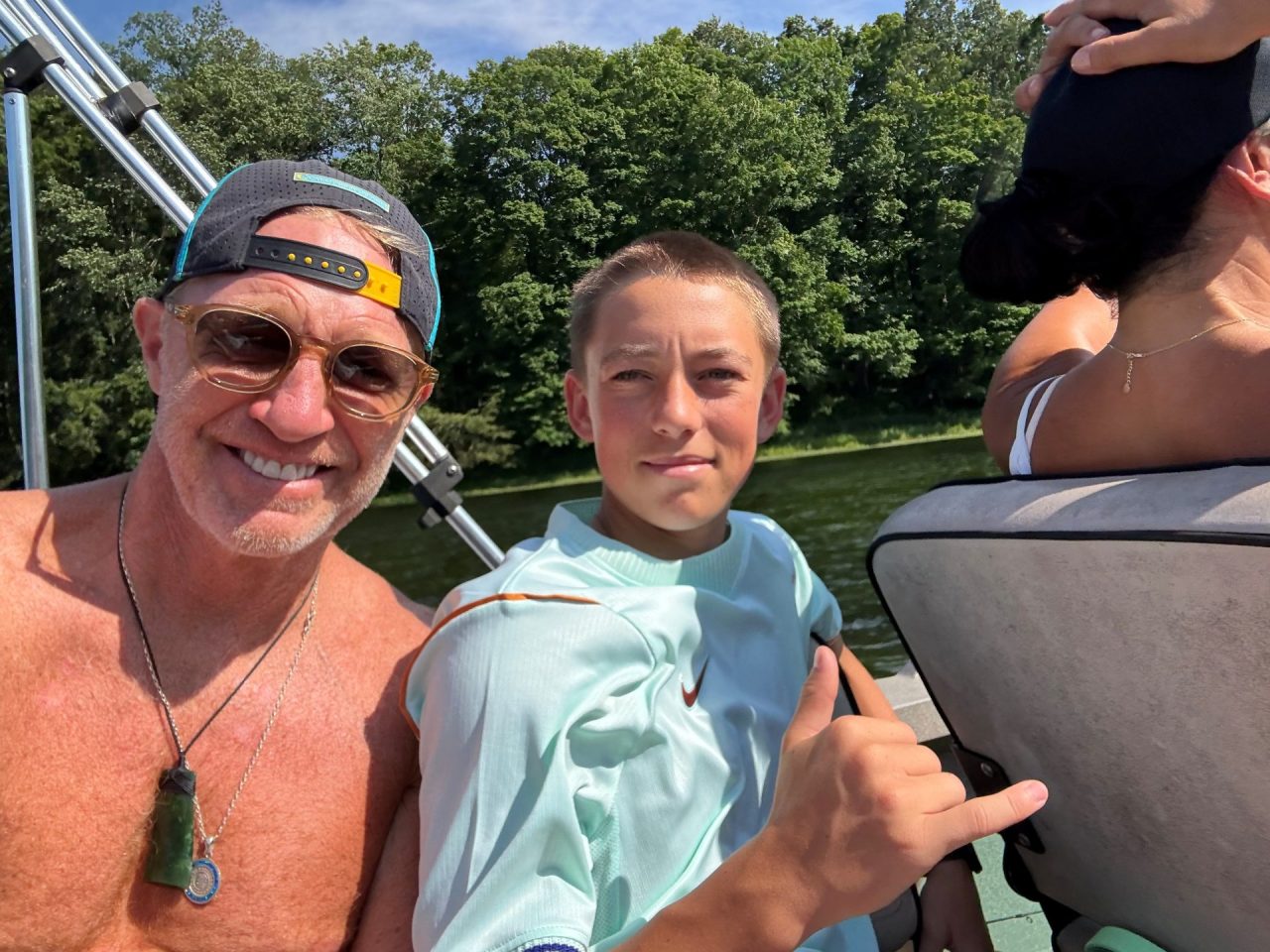 Three people on a boat posing for a selfie on a sunny day, with a man wearing a cap and sunglasses, a boy in a light blue shirt, and a woman facing away.