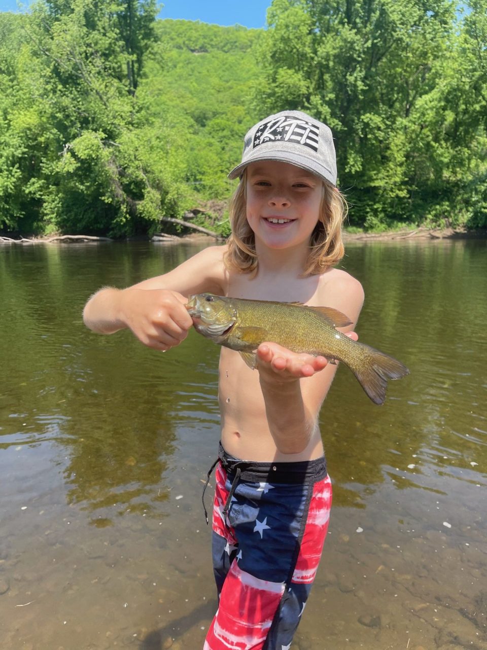 Smiling child in a gray cap holds a small fish over a river with green trees in the background on a sunny day.