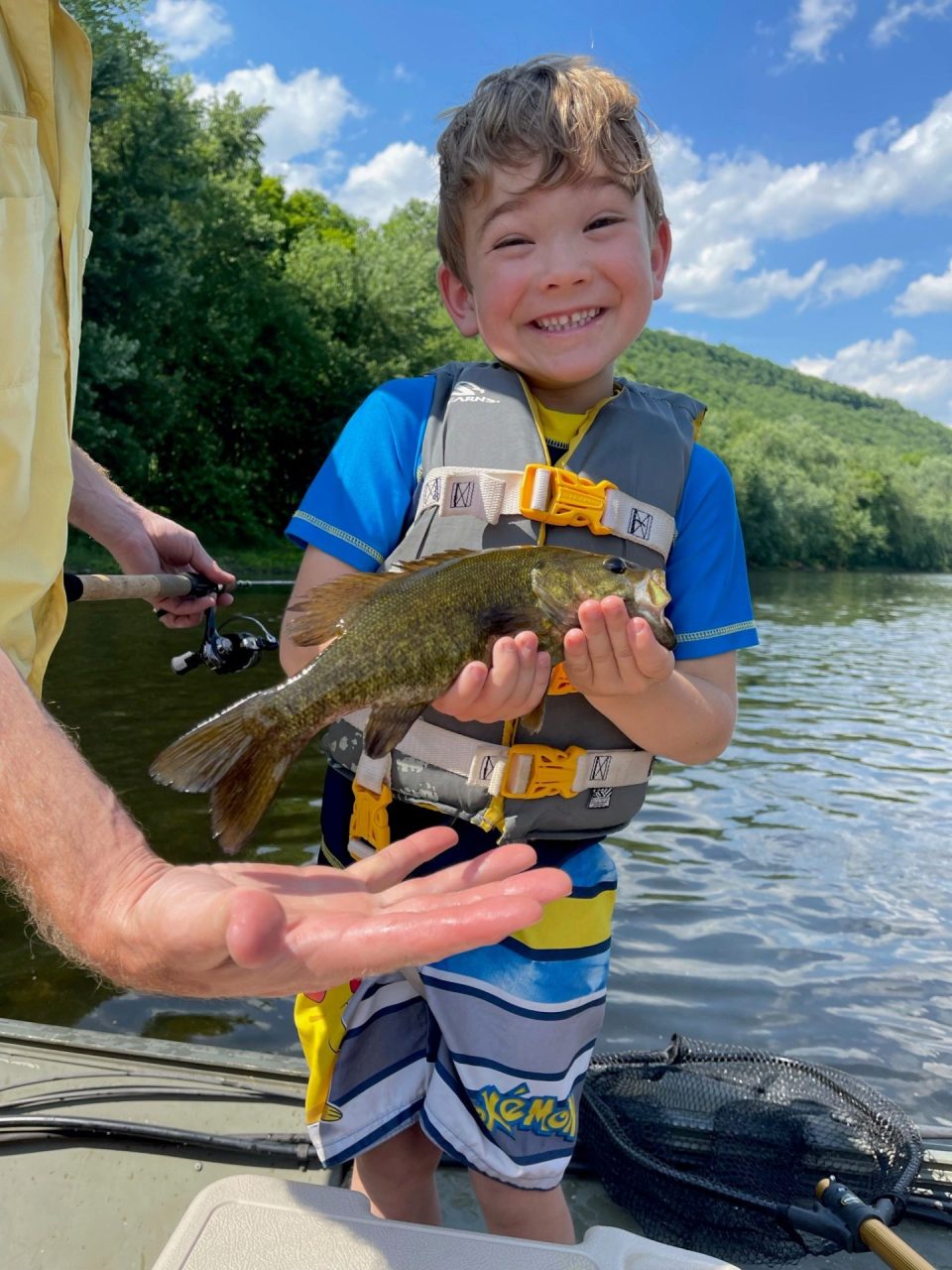 Happy young boy in a life jacket holds up a fish on a boat, blue sky and trees in the background.