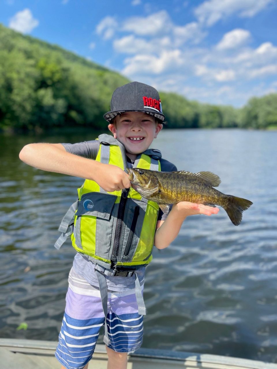 Smiling child in a life vest holds a fish on a lake with blue sky and trees in the background.