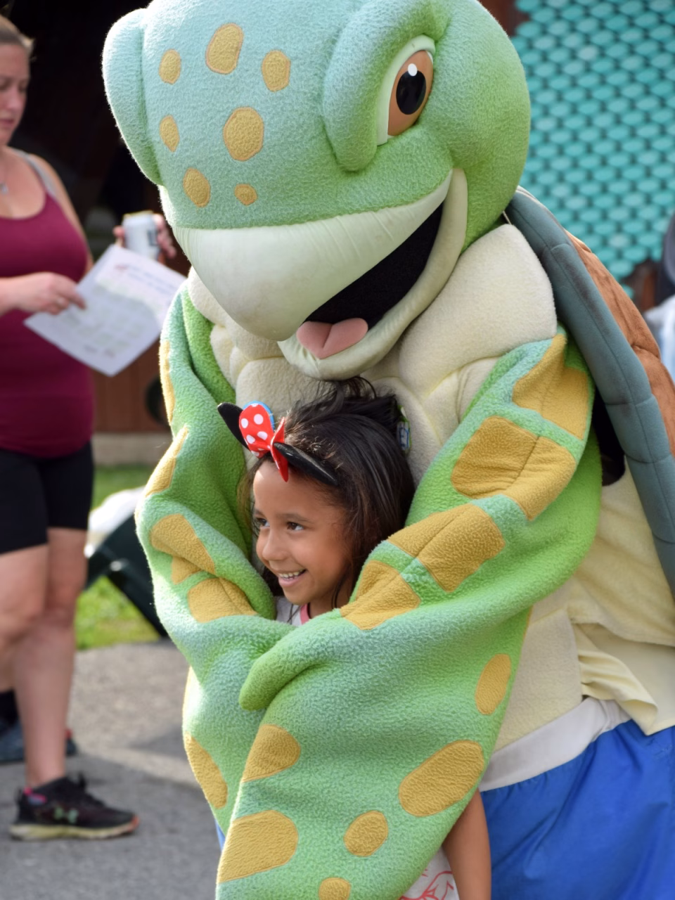 A young girl smiles as a green turtle mascot hugs her from behind at an outdoor event, with a woman in the background holding papers nearby.